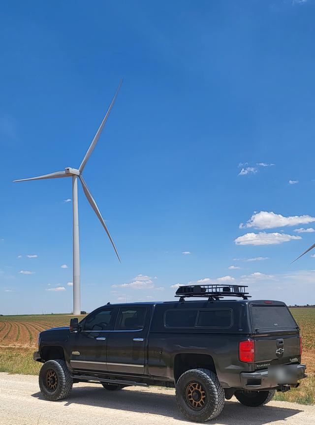 A black overland pickup parked on a dirt road beside a wind turbine and open farmland