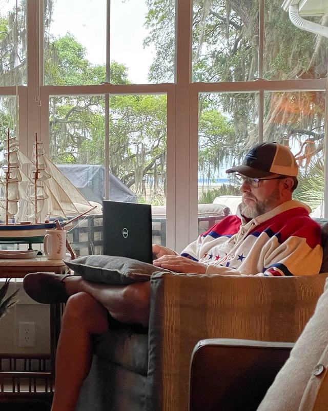 Shane Yerkes writing on a laptop in a sunroom, live oaks and Spanish moss through the windows behind him